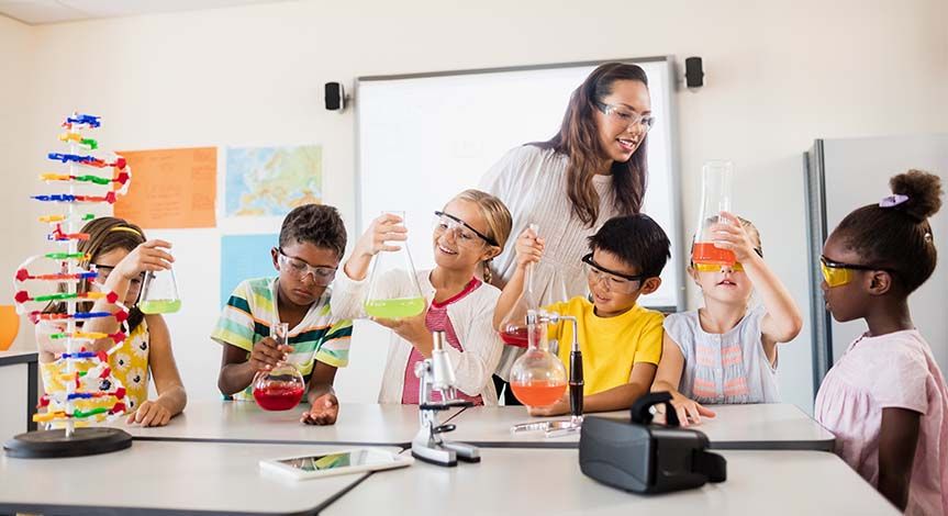 6 students looking at beakers with liquid in them.  Teacher standing over top supervising.  Also on the table is a microscope