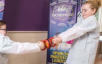 A boy and a girl in mad science lab coats playing with 2 magnets that are stuck together